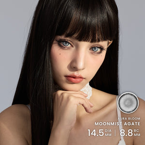 Close-up portrait of a woman with long dark hair and blue-gray eyes wearing colored contact lenses, with product text reading Aura Bloom Moonmist Agate and size details in the lower right.
