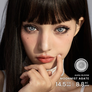 Close-up portrait of a woman wearing large blue-gray contact lenses, with text on the right reading 'AURA BLOOM MOONMIST AGATE 14.5 DIA MM | 8.8 Base curve