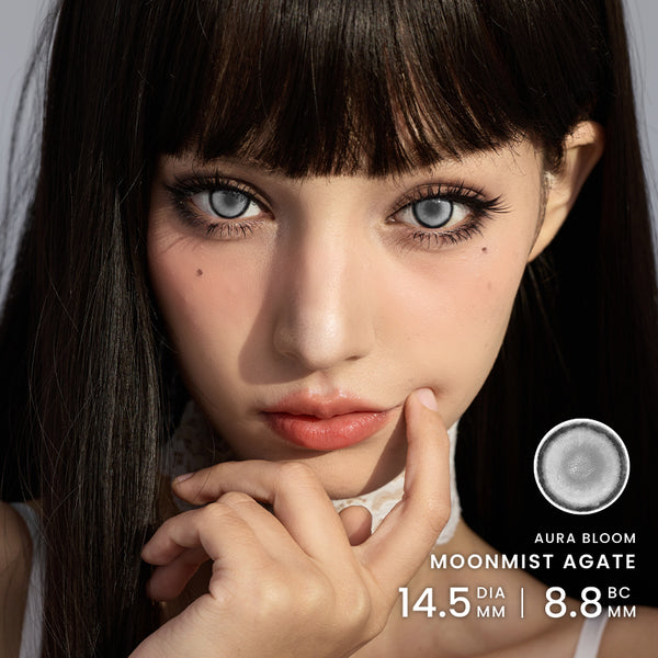 Close-up portrait of a woman wearing large blue-gray contact lenses, with text on the right reading 'AURA BLOOM MOONMIST AGATE 14.5 DIA MM | 8.8 Base curve